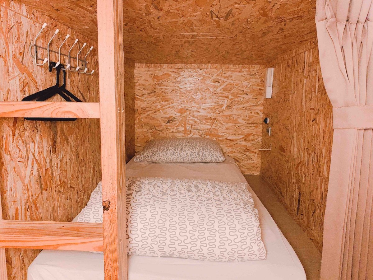A compact sleeping area is presented, featuring a spacious bed dressed in neutral bedding. Wooden panels provide a natural ambiance, while a curtain can be seen, allowing for privacy. Shelves with hangers are positioned nearby, contributing to the efficient use of space.