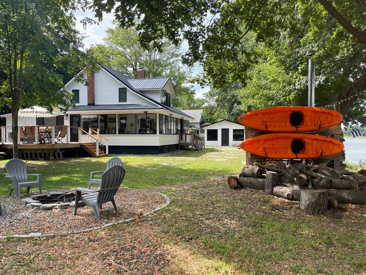 A grassy area features stacked logs and a fire pit surrounded by four comfortable chairs. Two kayaks in vibrant orange are positioned nearby. The two-story house, with large windows and a covered porch, offers views of the river through surrounding trees.