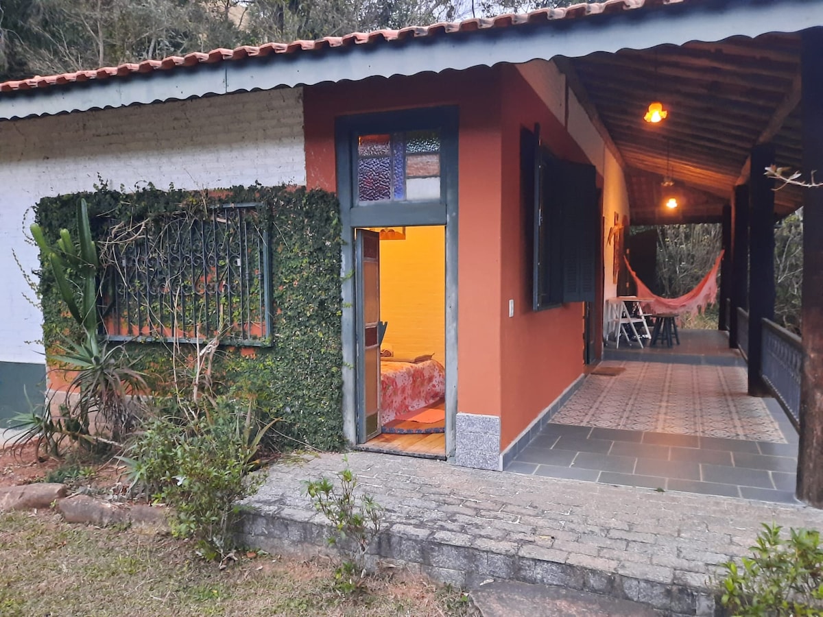 The exterior of a charming house is showcased, featuring a vibrant orange wall complemented by decorative green foliage. A cozy porch with a hammock invites relaxation, while a glimpse is provided into the warm interior through the open door, revealing a comfortable sleeping area.