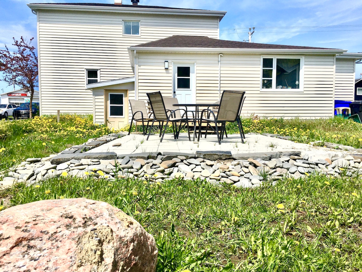 A well-maintained outdoor area features a stone-lined seating space equipped with four chairs and a small table. The exterior of the house is visible, showcasing light-colored siding and several windows, with green grass and dandelions surrounding the space.