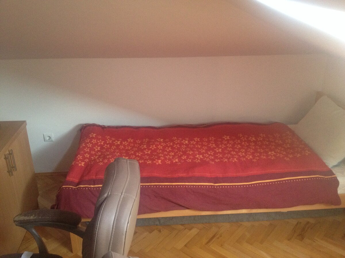 A simple sleeping area features a narrow bed adorned with a red and gold patterned blanket. A pale-colored wall provides a neutral backdrop, while a light-colored wooden cabinet is positioned nearby. A chair with a cushioned seat is also visible in the corner.