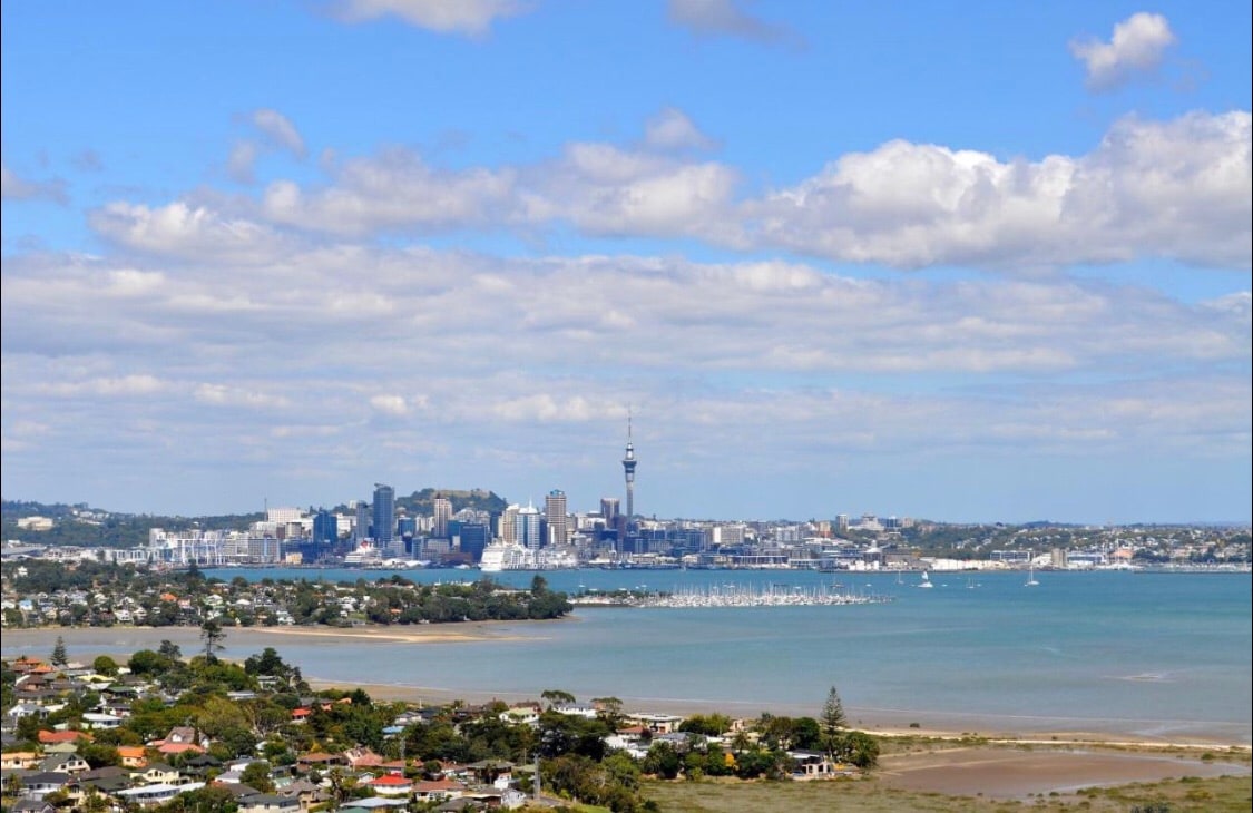 A panoramic view showcases the Auckland city skyline, featuring the Sky Tower among other buildings. The foreground displays coastal waters with boats, while the surrounding landscape includes residential areas and lush greenery under a partly cloudy sky.