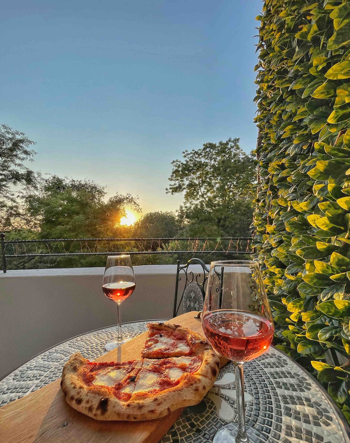 A small table is set on a balcony, featuring a freshly baked pizza on a wooden board and two glasses of rose wine. The sun is setting in the background, casting a warm glow over the greenery surrounding the space.