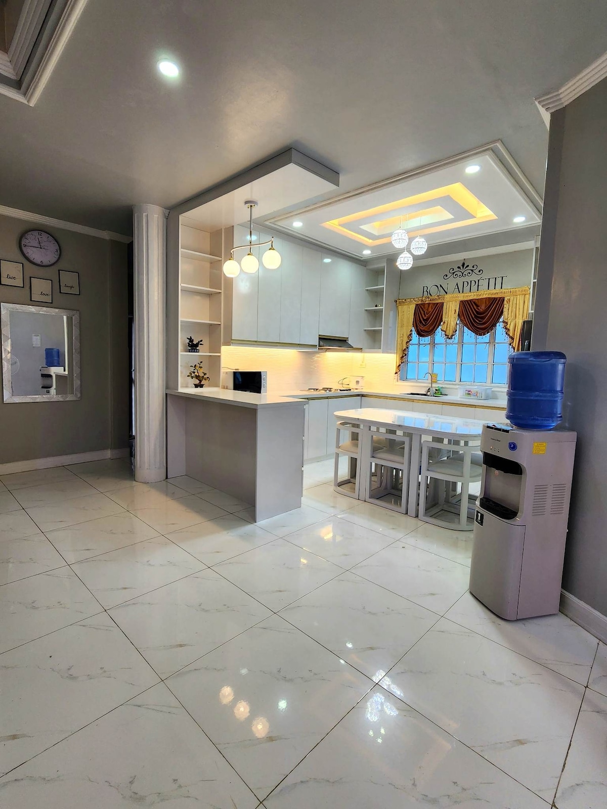 A modern kitchen area is depicted, featuring sleek cabinetry and a central island with seating. Bright overhead lights illuminate the space, while a water dispenser stands nearby. Large windows allow natural light to fill the room, creating a welcoming atmosphere.