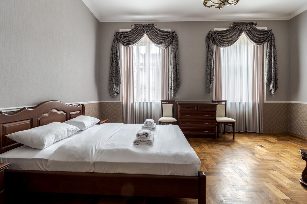 A spacious bedroom features a wooden bed with neatly arranged linens. Two large windows are adorned with elegant drapes, allowing natural light to brighten the room. A wooden dresser and two chairs complement the warm tones of the hardwood floor.