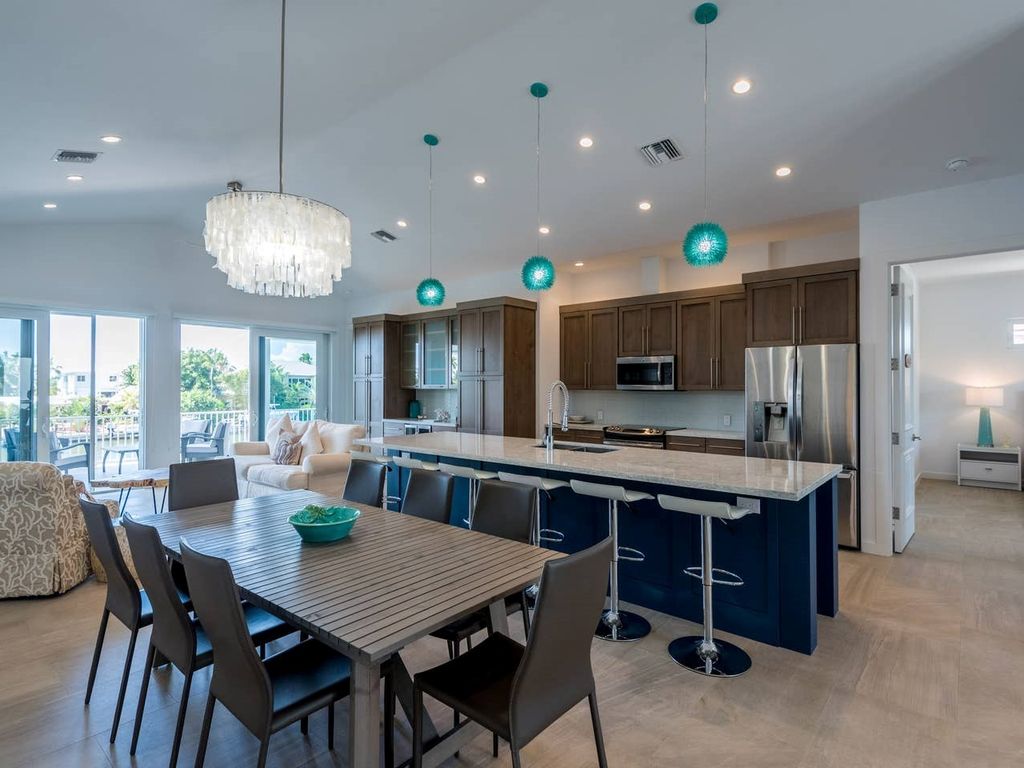 A modern kitchen and dining area feature a large wooden table surrounded by chairs. Pendant lights hang above the oversized island, which includes bar seating. Bright windows offer views of the outdoors, while cabinets and appliances showcase a contemporary design.