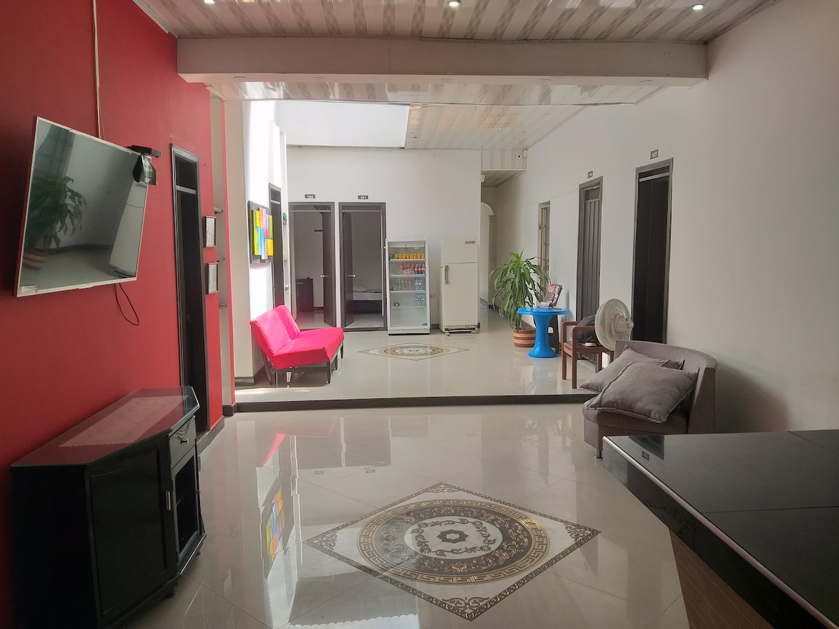 A shared common area features sleek decor with a mix of vibrant red and neutral tones. A modern flat-screen TV is mounted on the wall, with a pink couch and a gray sofa offering seating. A large, decorative tile adds visual interest to the floor.