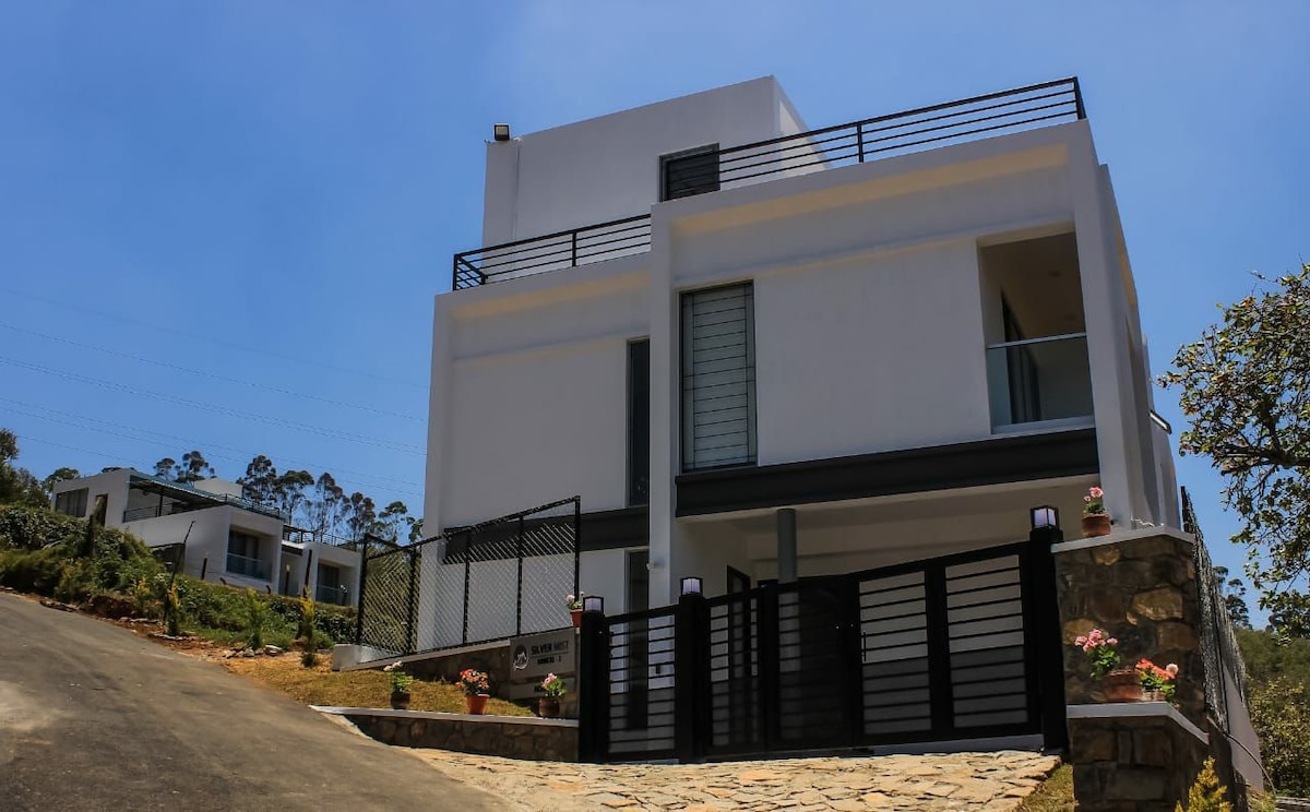A modern three-story villa stands prominently with a sleek facade, showcasing large windows and a neutral color palette. A stone pathway leads to the gated entrance, flanked by flowering plants. Lush greenery surrounds the property, creating a serene environment.