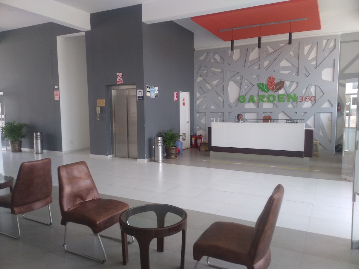 A modern lobby area is showcased, featuring a reception desk adorned with the 'GARDEN 360' logo. Brown chairs and a glass-top table provide seating. Large windows allow natural light to fill the space, while decorative plants add a touch of greenery.