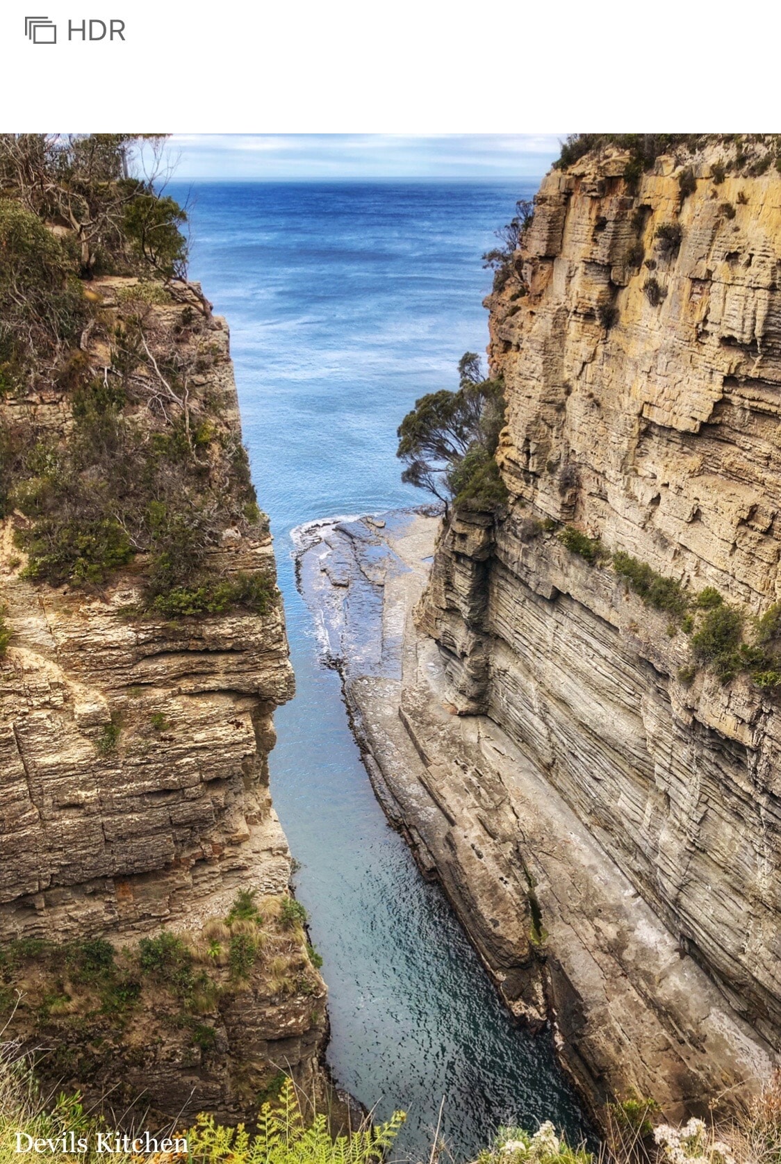A dramatic coastal scene captures steep cliff faces adorned with textured rock layers, flanking a narrow waterway. The blue sea gently glistens in the sunlight, while scattered greenery accentuates the rugged terrain, creating a serene atmosphere against the expansive sky.