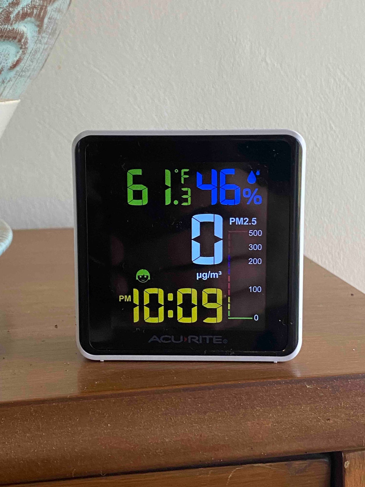 A digital display shows indoor temperature at 61.3°F and humidity at 46%. Additional readings include PM2.5 values and the current time, 10:09 PM. The device is placed on a wooden surface, featuring a simple design with bright, easy-to-read graphics.