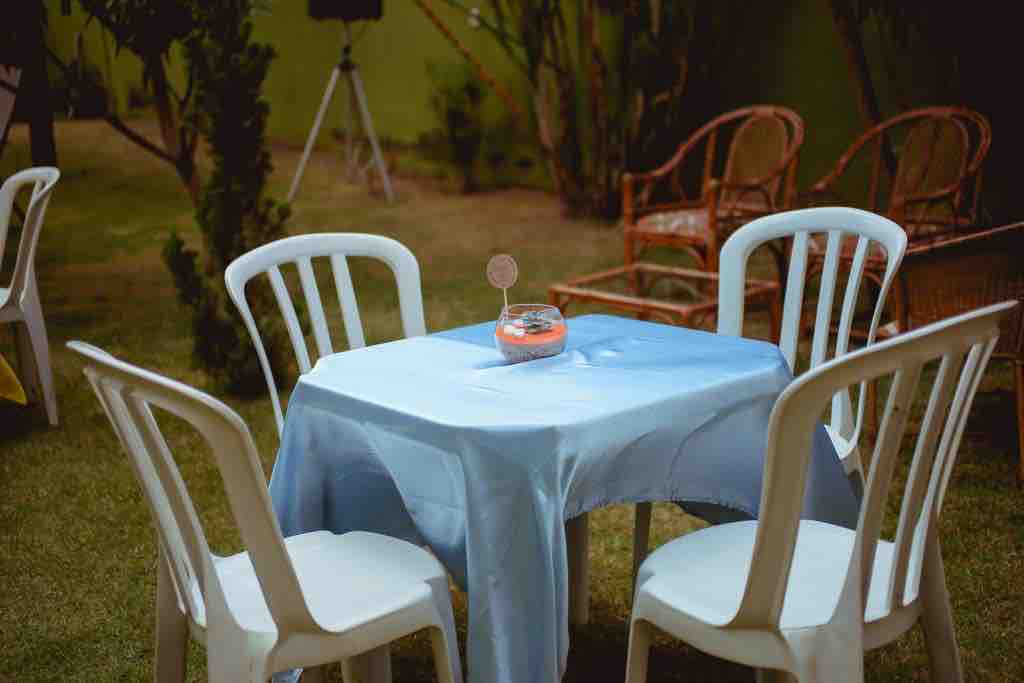 A small outdoor table is set with a light blue tablecloth and a decorative item at its center. Four white chairs surround the table, positioned on a grassy area. In the background, a seating area with wicker chairs is partially visible among greenery.