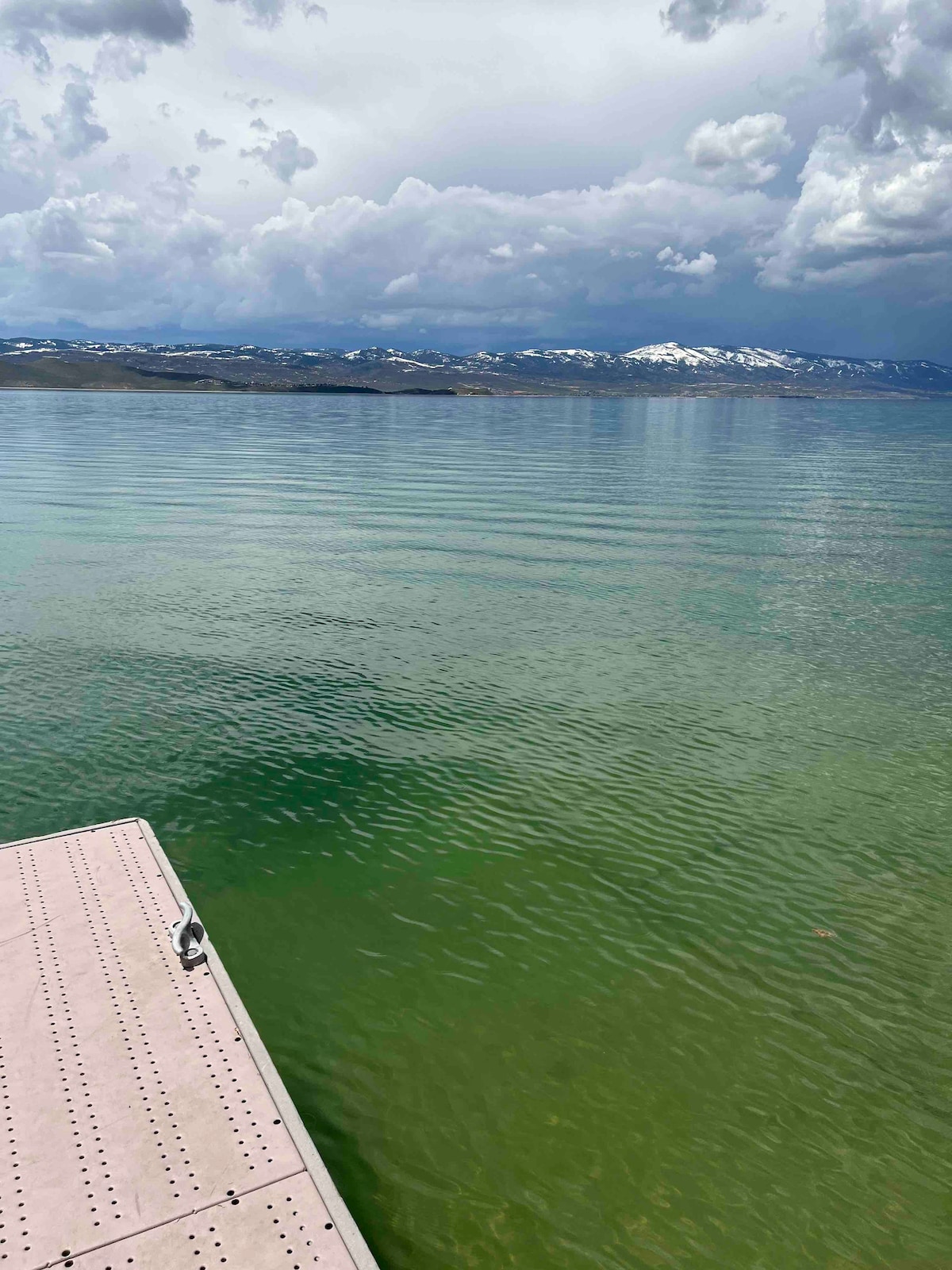 A view of a tranquil lake is captured, with calm waters reflecting the clouds overhead. In the foreground, a wooden dock extends into the water, while distant mountains create a scenic backdrop, partially covered in snow.