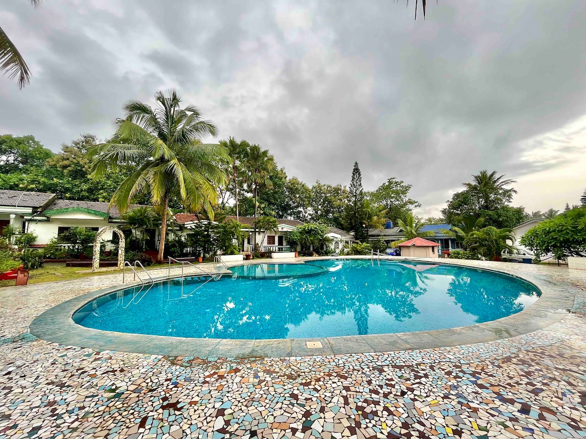A serene shared swimming pool is set within a lush garden, surrounded by palm trees and well-maintained cottages. The tranquil water reflects the cloudy sky, inviting guests to relax by its mosaic-tiled edges.