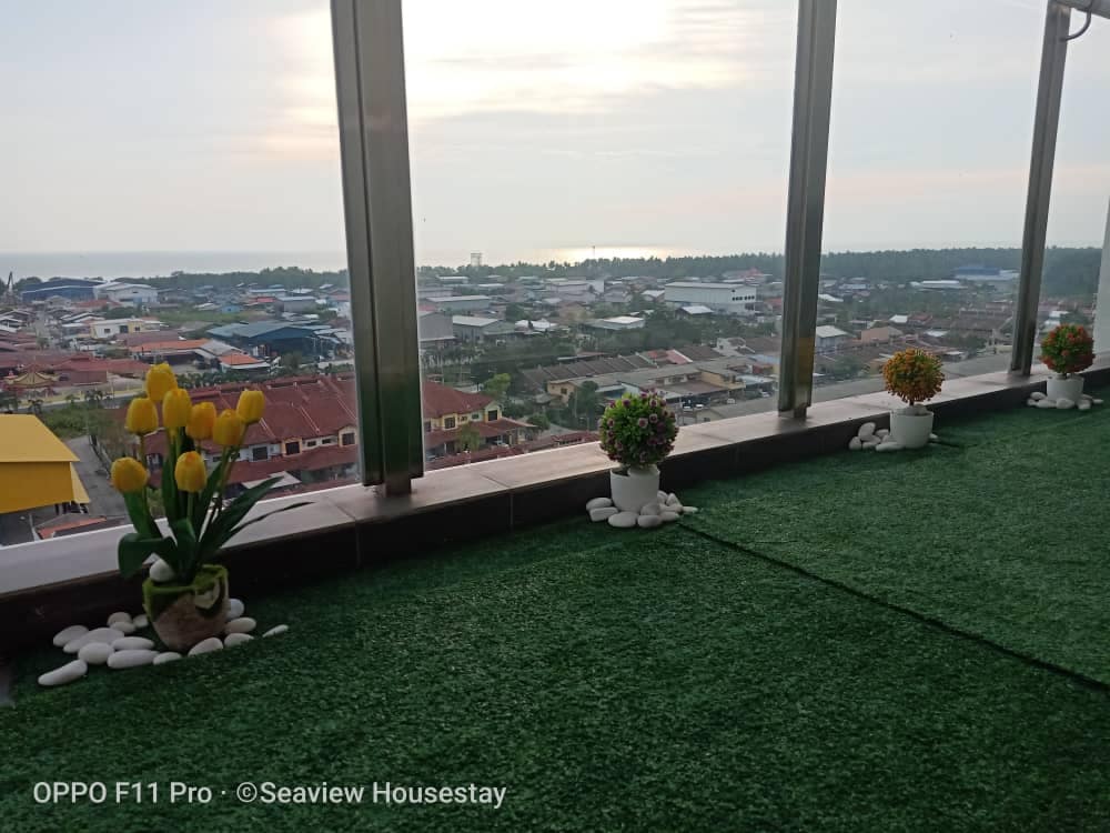A spacious balcony area features artificial grass, bordered by potted plants with colorful blooms. Smooth white stones accent the space, creating a serene atmosphere. In the background, a panoramic view of the ocean and distant rooftops is visible under a softly lit sky.