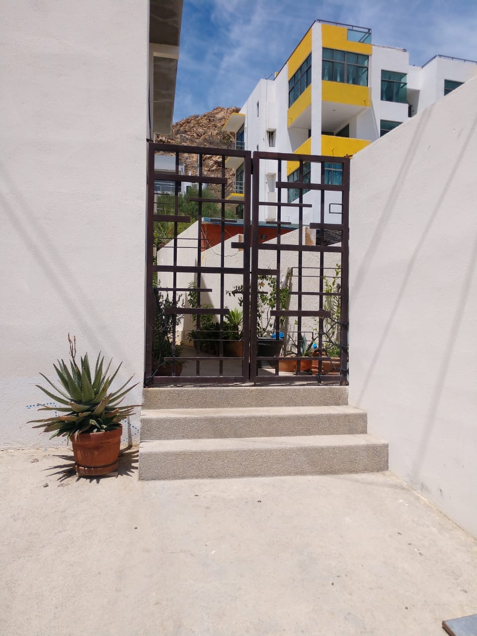 An entrance gate is framed by beige walls, inviting guests into a serene outdoor space. Steps lead up to the gate, while a potted plant sits beside the entrance, adding a touch of greenery to the setting. The background reveals a glimpse of buildings and a clear blue sky.