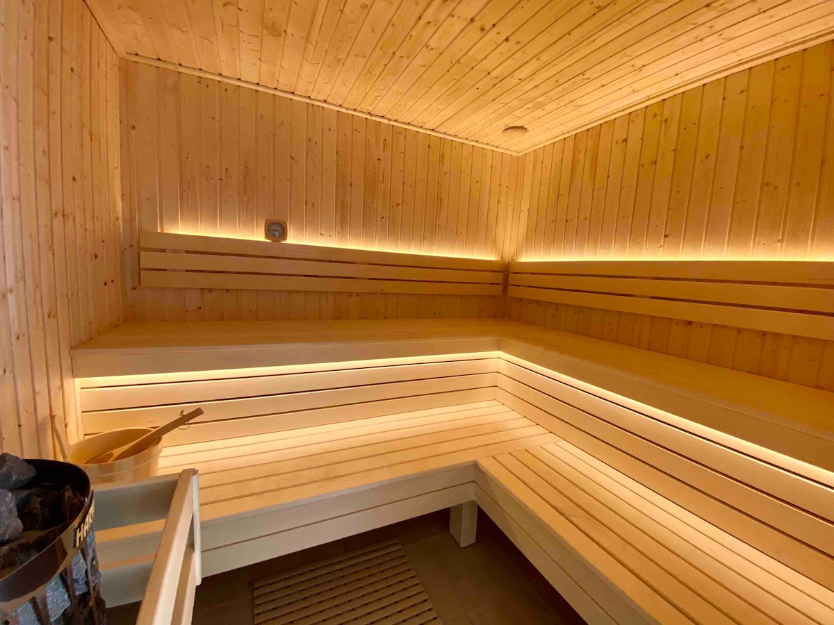 The sauna features warm wooden paneling and ambient lighting, creating a soothing environment. Bench seating is arranged along the walls, providing ample space for relaxation. A small heating unit with stones is visible, enhancing the tranquil atmosphere.