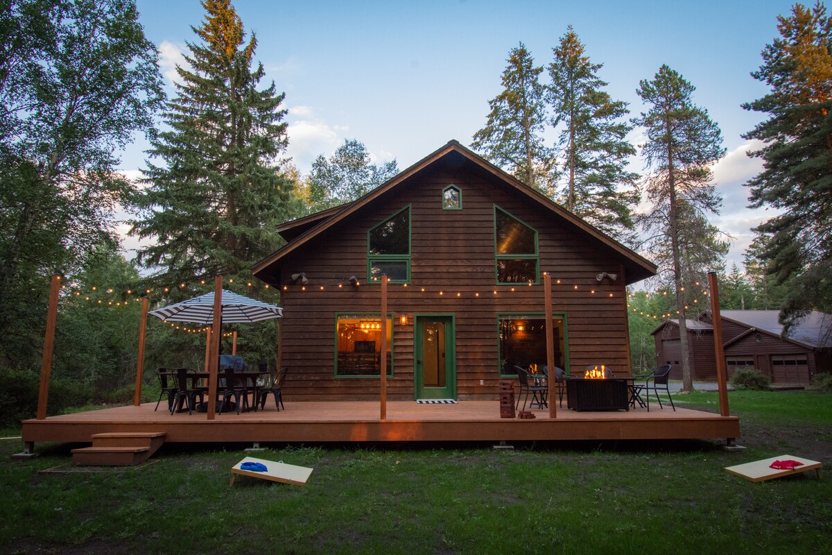 A spacious cabin is seen surrounded by tall trees, with a large wrap-around deck hosting outdoor seating. Soft lights adorn the deck, creating a welcoming atmosphere. The front door is flanked by large windows, providing views of the natural surroundings.