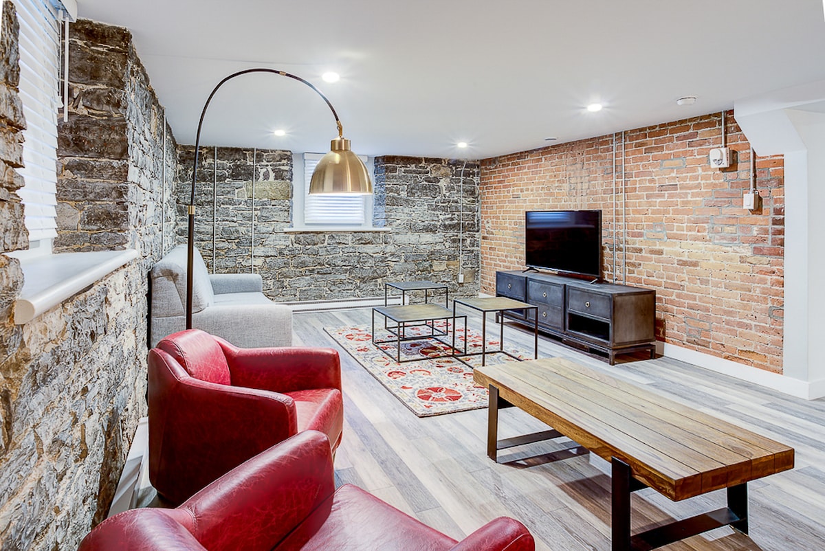 A spacious living area features exposed stone and brick walls, providing a rustic charm. A comfortable gray sofa and two red armchairs create a cozy sitting arrangement around a wooden coffee table. A wall-mounted television is positioned opposite, enhancing the modern aesthetic.