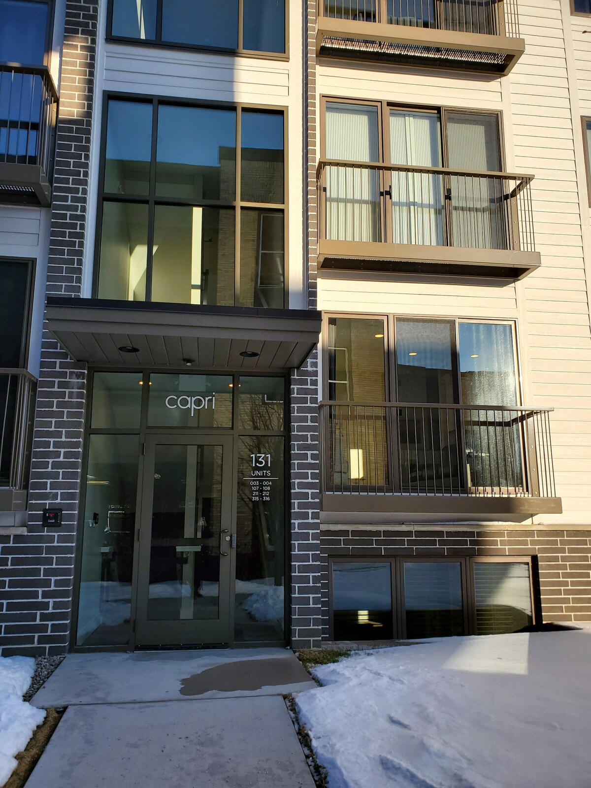 The building entrance is featured, showcasing modern architecture with large windows and a glass door labeled 'capri.' Balconies can be seen on the upper floors, and snow covers the ground, indicating a winter setting.