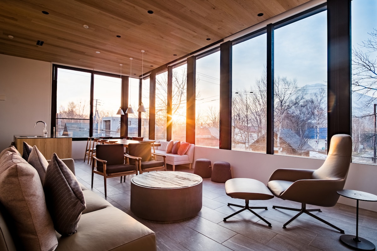 A spacious living area bathed in natural light features floor-to-ceiling windows that frame a sunset view. Modern furniture, including a cozy sofa and armchair, is arranged around a circular coffee table. Wooden accents are present in the ceiling and floor details.