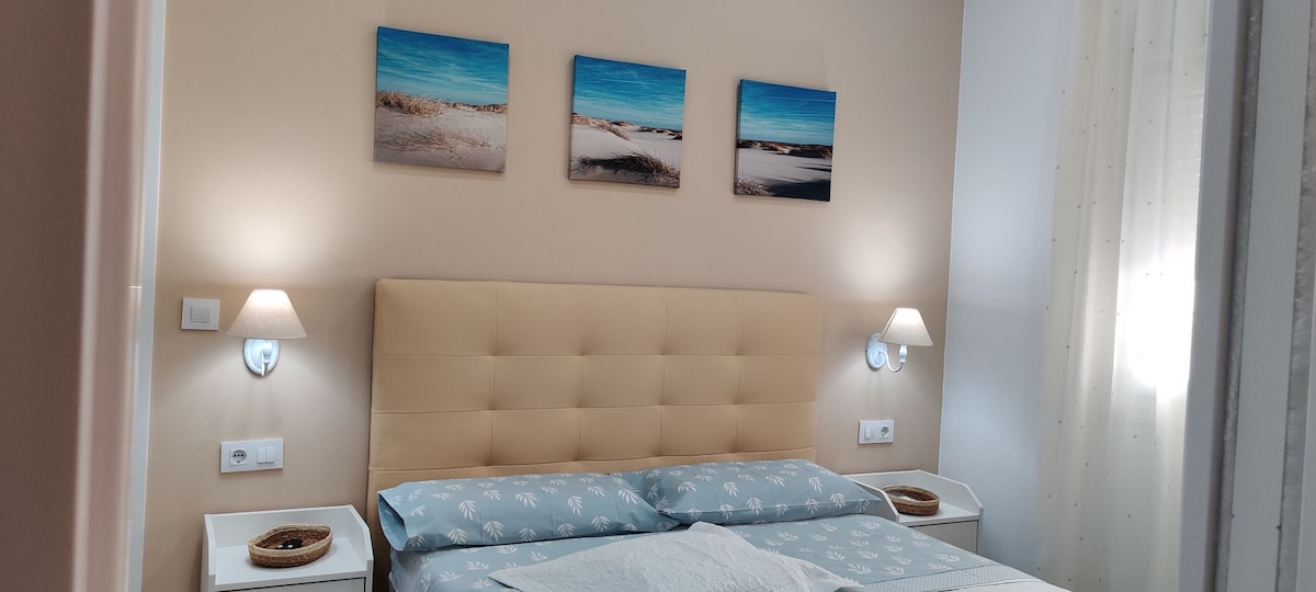 The bedroom features a spacious bed with a soft headboard, complemented by bedside tables and lamps. Three landscape artworks hang above the bed, while natural light filters through sheer curtains, creating a calm atmosphere.