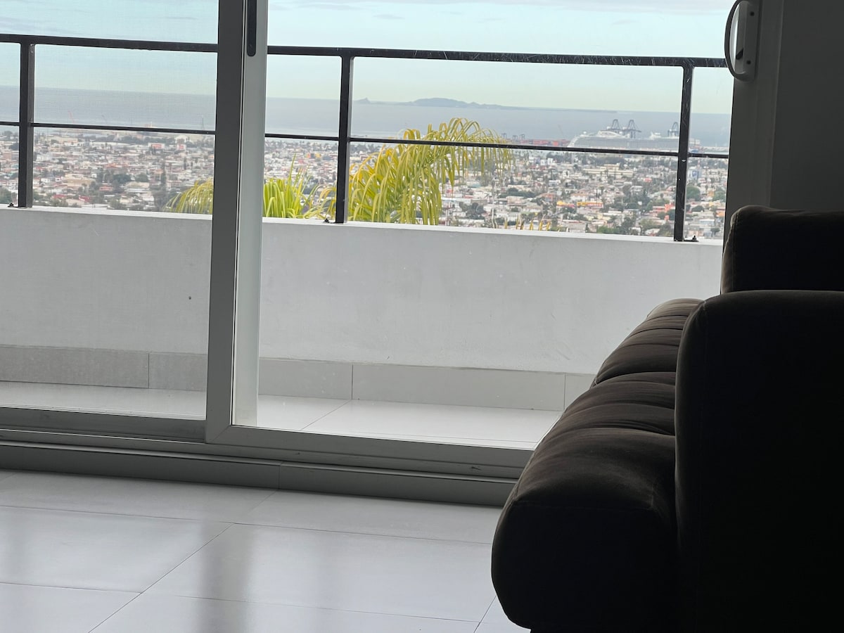 A spacious living area is visible, featuring a comfortable brown sofa. Large sliding glass doors open to a balcony, revealing a panoramic view of the city and coastline beyond. The interior is bathed in natural light, highlighting the clean lines of the space.
