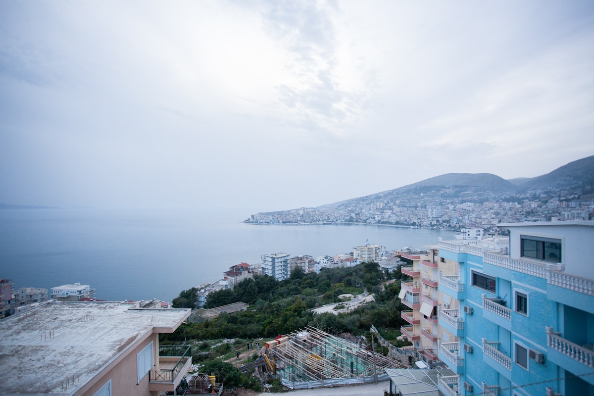 A panoramic view of the tranquil seascape featuring the coastline of Corfu Island and Saranda. The image captures the soft hues of the sky reflecting over the water, with buildings lining the shore and a distant mountainous backdrop.