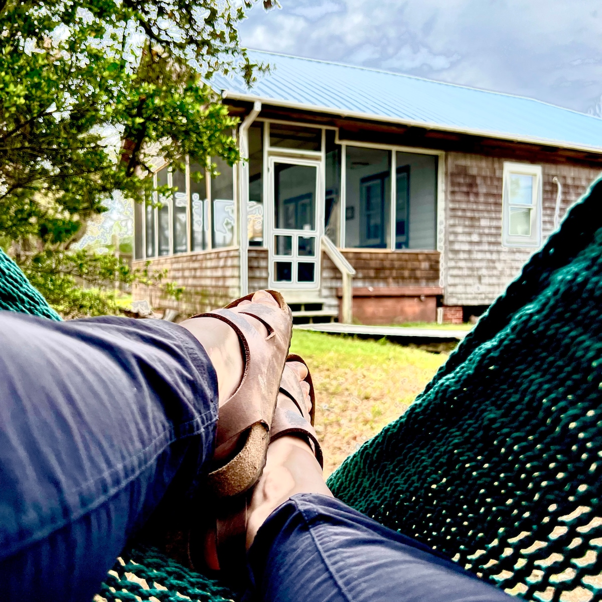 A hammock rests in the foreground, with relaxed feet visible in sandals. In the background, the cottage is presented, showcasing its wooden exterior and screened porch. Surrounding greenery adds to the serene atmosphere of the outdoor space.