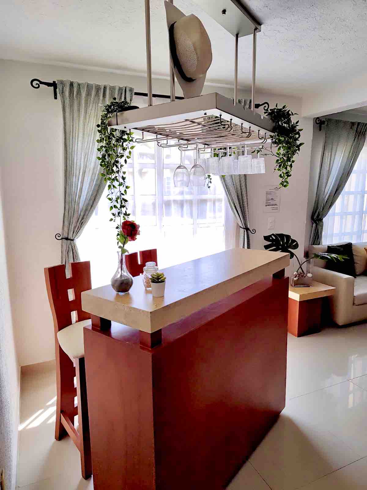 A stylish kitchen bar area features a countertop with bar stools at one side. Above, hanging glass holders and decorative plants add a touch of greenery. Natural light floods through large windows, complemented by soft drapes and a cozy seating area nearby.