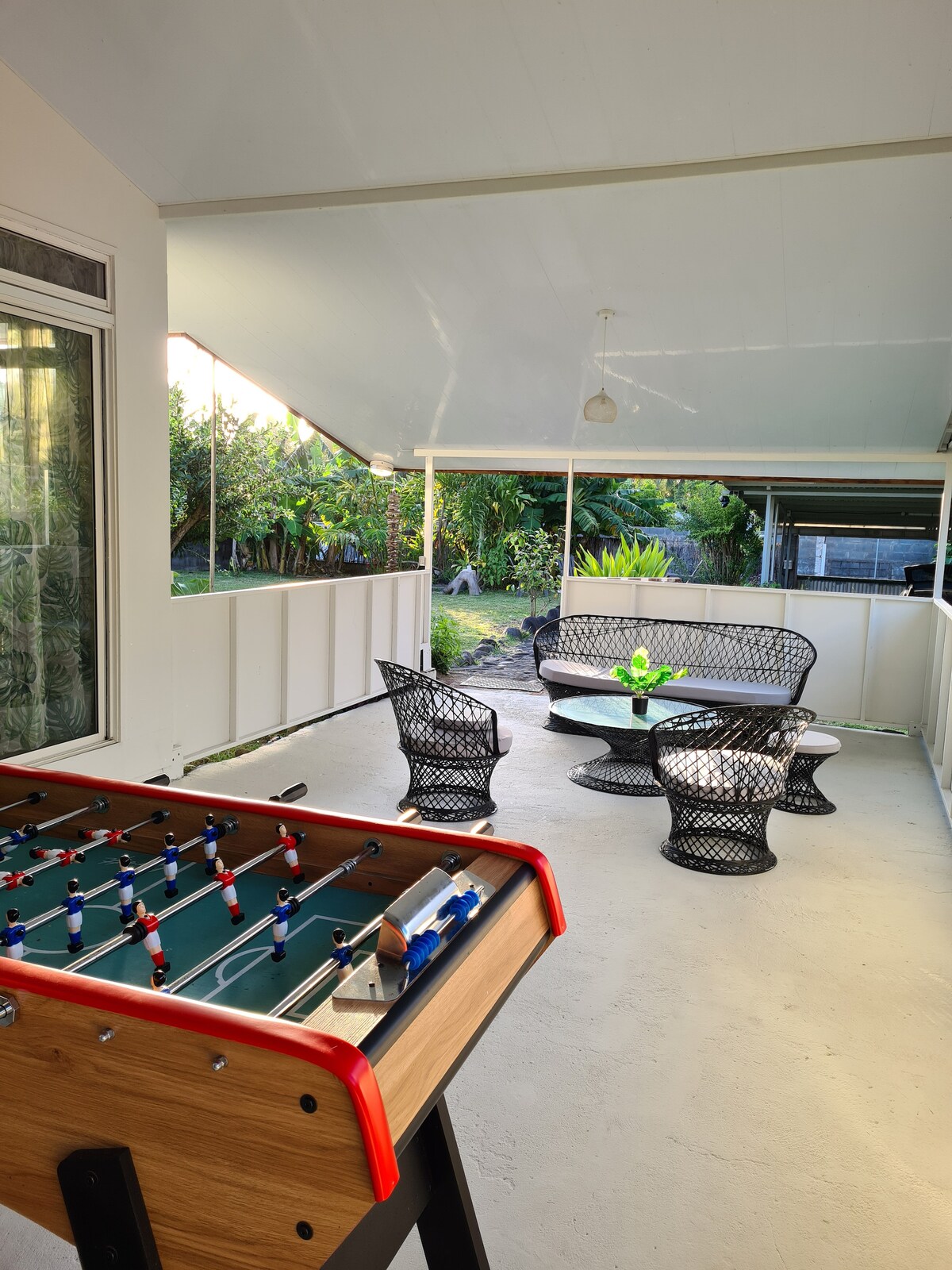 A covered terrace is showcased, featuring a foosball table in the foreground. In the background, a seating area consists of a round table surrounded by four woven chairs, creating a cozy spot for gatherings. Lush greenery is visible through the open space, enhancing the tranquil setting.