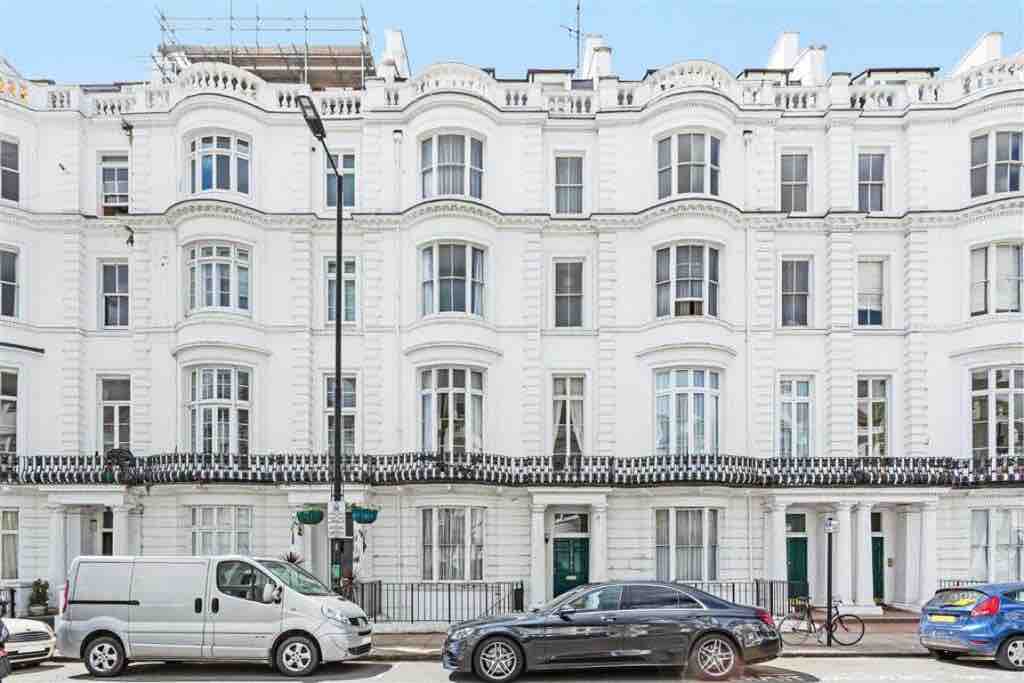 The elegant building façade features a classic white exterior with ornate architectural details. Large windows are aligned symmetrically, and ornamental wrought iron railings adorn the balconies. The street view showcases parked cars and well-maintained sidewalks, contributing to the urban appeal of the setting.
