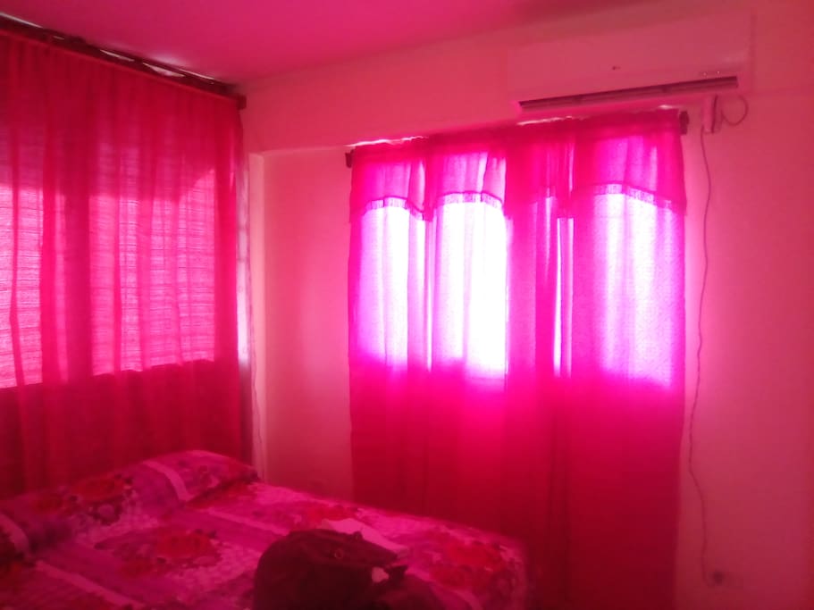 Un cuarto con aire acondicionado y cortinas rosadas. Tiene muy buena iluminación. El baño es intercalado.