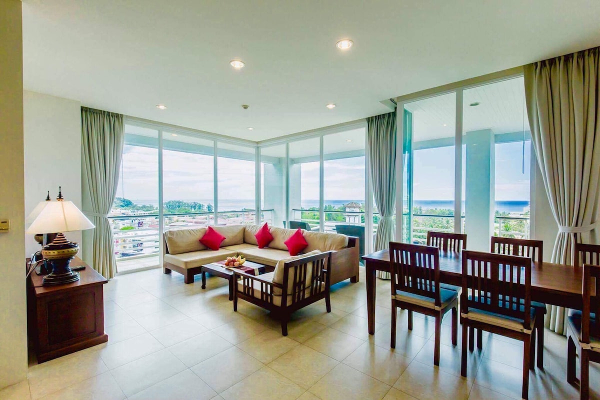 A bright living area features large windows offering expansive sea views. A comfortable sofa is adorned with decorative cushions, facing a wooden coffee table. A dining table with chairs is positioned nearby, creating an inviting space for relaxation and meals.