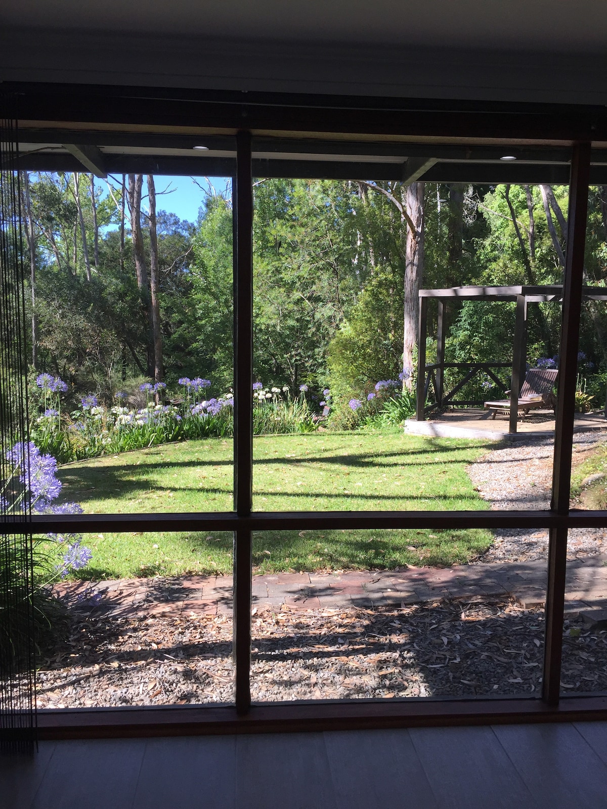 A view of the garden is framed by large glass doors, showcasing lush greenery and native flowers. A shaded gazebo is visible, providing a peaceful spot amidst the natural surroundings. Light filters through the leaves, creating a serene atmosphere within the studio.