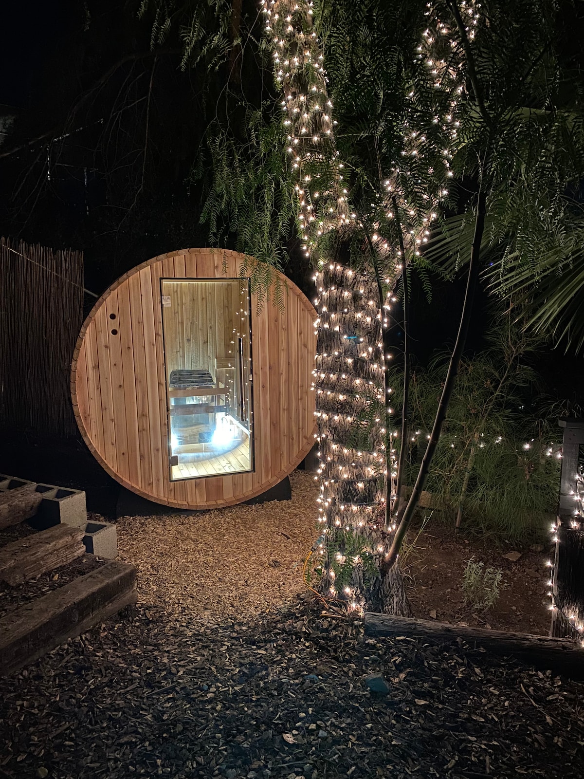 A wooden barrel sauna is illuminated at night, surrounded by a softly lit tree adorned with string lights. The entrance of the sauna reveals a serene interior, inviting relaxation amidst the natural setting.
