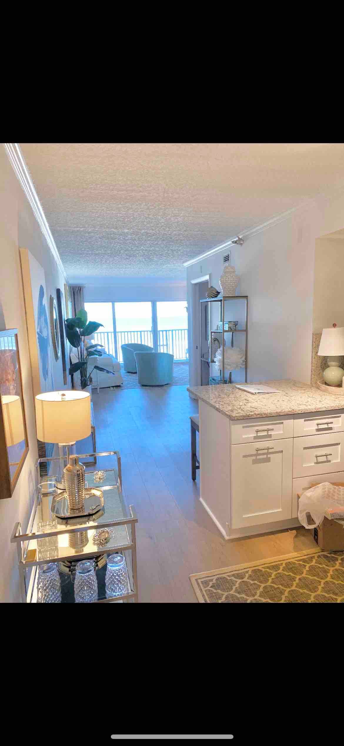 A bright and airy living space features a light-colored floor and soft neutral walls, creating a calming environment. Natural light floods in through large windows at the end of the room, highlighting a dining area with a stylish table and two comfortable chairs. Decorative elements include a lamp and a shimmering cart.