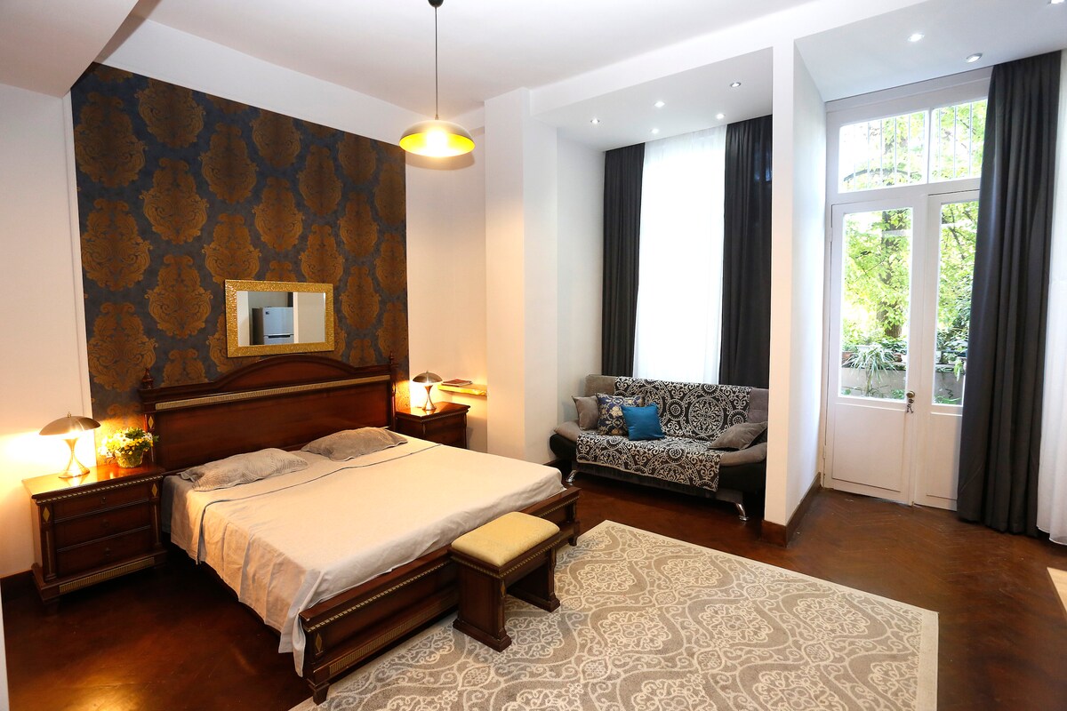 A spacious bedroom features a comfortable bed with a light-colored bedspread, flanked by wooden nightstands. A large mirror hangs above the bed, reflecting natural light from the nearby windows, which are adorned with dark curtains. A cozy seating area includes a patterned sofa.
