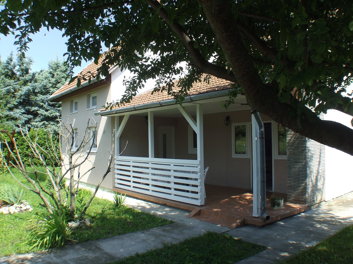 A renovated standalone vacation house is set in a peaceful street, featuring a front porch with white railings. Lush green grass surrounds the property, complemented by trees and shrubs. The exterior is designed with a combination of light-colored walls and a tiled roof.