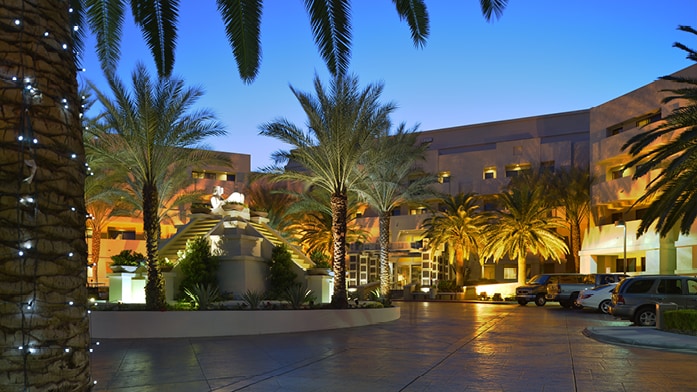 The entrance area is surrounded by palm trees and manicured landscaping, illuminated by soft lighting at dusk. A decorative fountain is centered in the foreground, with a spacious parking area visible to the right and welcoming architectural features of the resort in the background.