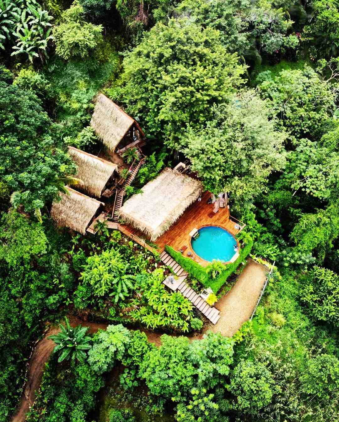 An aerial view captures three charming bungalows nestled among lush greenery. A round pool is centered within the property, surrounded by a wooden deck and tropical plants, creating a serene outdoor oasis seamlessly integrated with nature.
