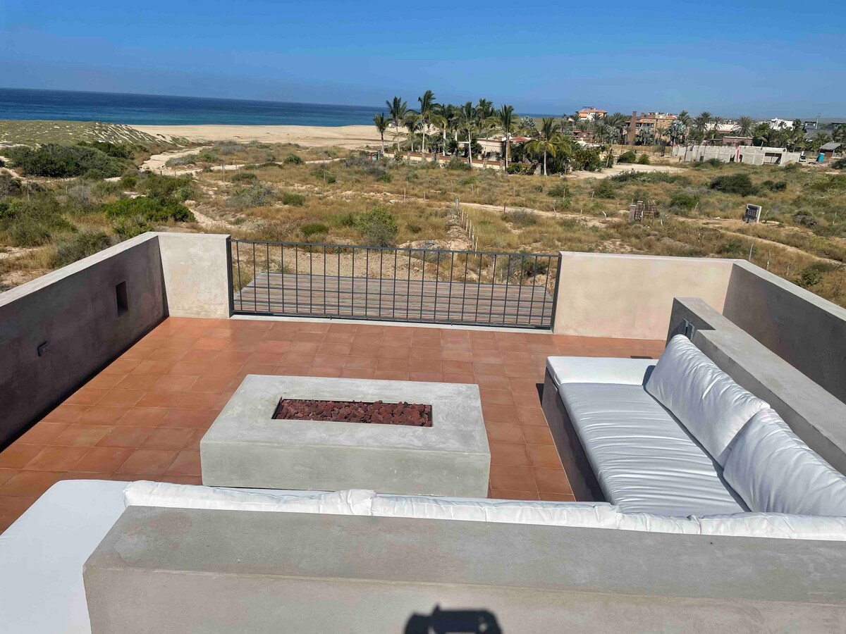 A spacious rooftop terrace is presented, featuring a comfortable u-shaped seating area with soft cushions. A modern fire pit sits at the center of the terrace. Views of the sandy beach and palm trees are visible in the distance, providing an inviting outdoor atmosphere.