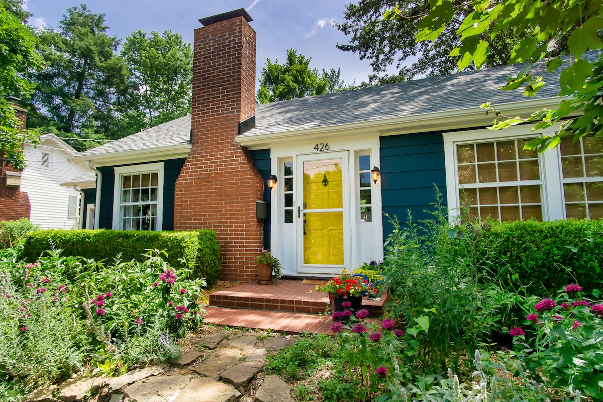 The charming exterior of the cottage is framed by vibrant seasonal blooms and well-maintained greenery. A yellow front door adds a pop of color, while a brick chimney rises against a backdrop of lush trees. The inviting pathway leads to the entrance, enhancing the home's curb appeal.