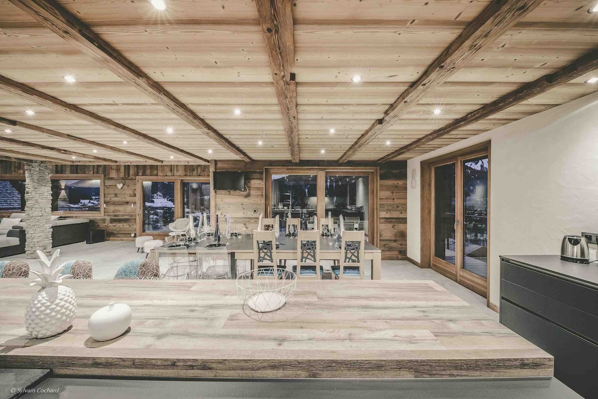 The interior space features wooden ceilings and walls, creating a warm ambiance. A large dining area is visible with multiple white chairs around a spacious wooden table. Floor-to-ceiling windows allow for mountain views and ample natural light, while a cozy seating area is seen in the background.