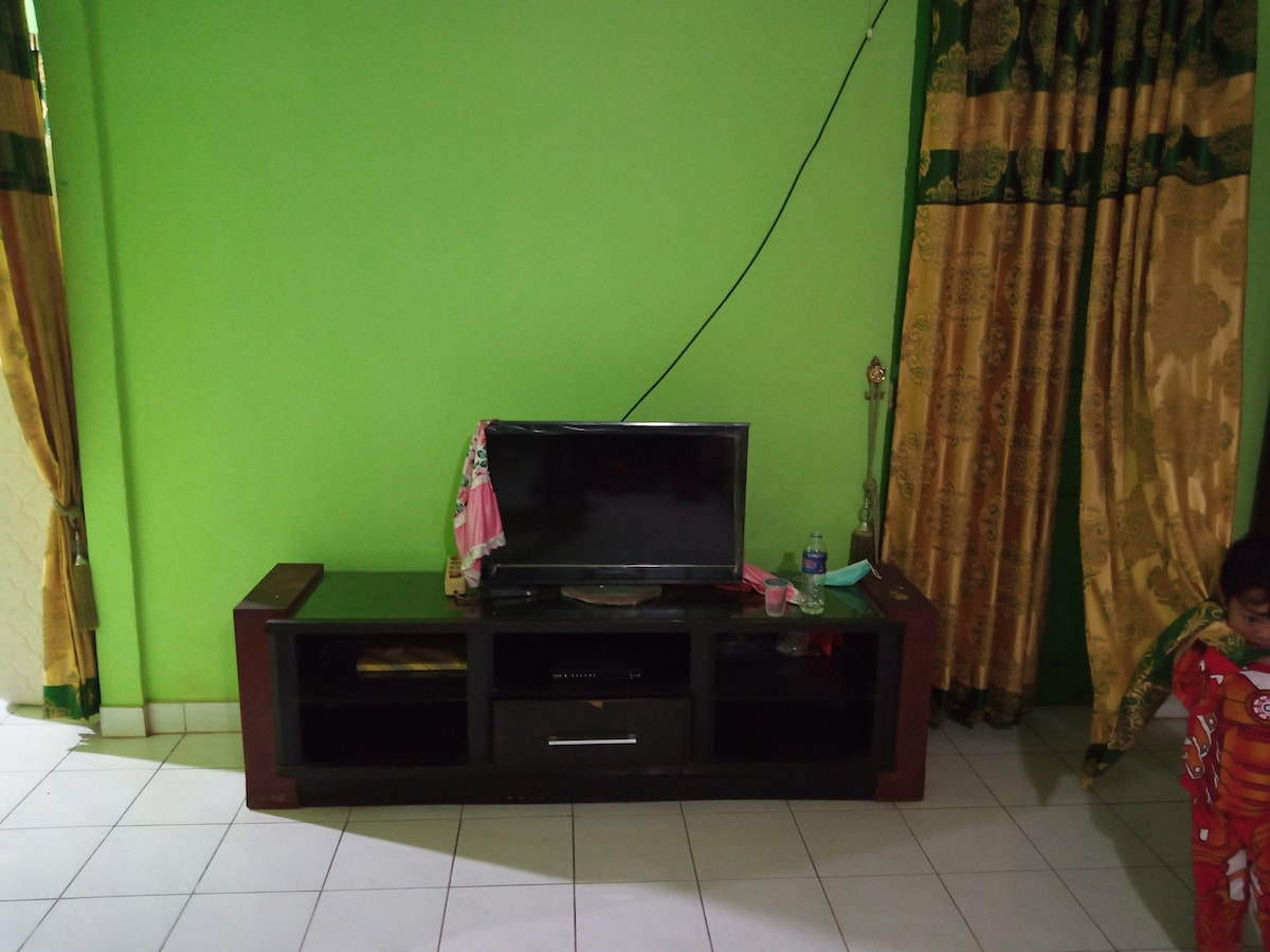 A living area is presented with a green accent wall, featuring a dark wooden TV stand that supports a flat-screen television. Gold and green curtains frame the space, while a few items are placed on the stand, including a water bottle and a small decorative piece.