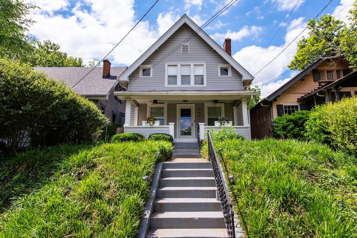 The exterior of the house, featuring a welcoming front porch, is surrounded by lush greenery. A staircase leads up to the entrance, framed by neatly maintained shrubs. The architectural design reflects a blend of classic and modern elements, complemented by a bright sky and scattered clouds.