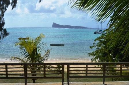 A serene view of the coastline features gentle waves lapping at the sandy shore. A small boat is anchored nearby, while a distant island silhouette is visible against the sky. Lush palm fronds frame the scene, adding to the tropical ambiance.