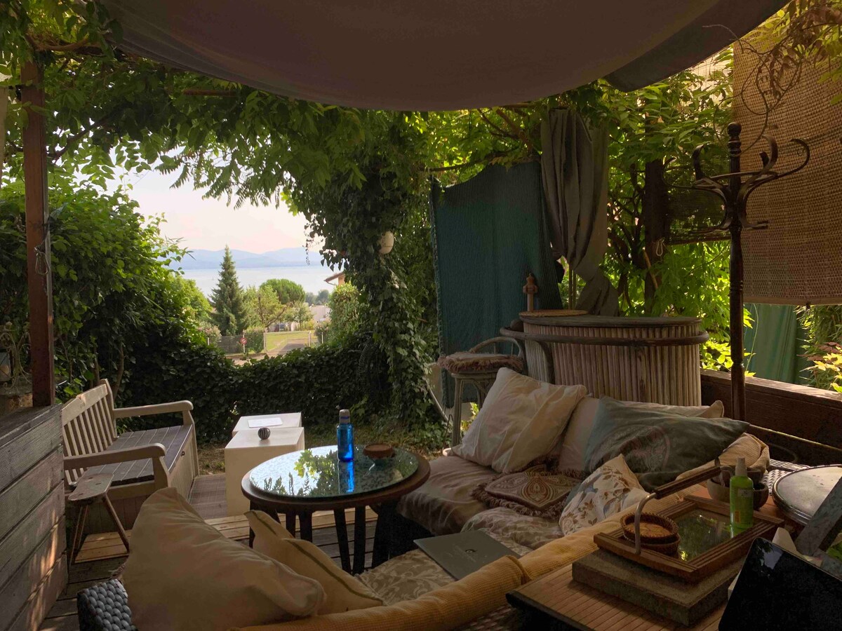 A shaded terrace area is surrounded by lush greenery, providing a serene atmosphere. Comfortable seating and a low table offer a cozy setting, with a glimpse of the lake visible beyond the foliage. Soft textiles and decorative elements create a welcoming space.