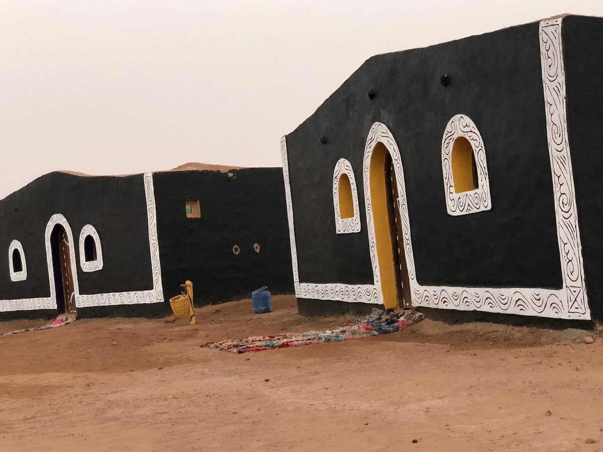 Adobe rooms are depicted in earthy tones, featuring curved white patterns along the edges. Each room is fitted with arched yellow windows that complement the rustic design, while colorful woven rugs are laid across the sandy ground, enhancing the traditional aesthetic of the desert camp.