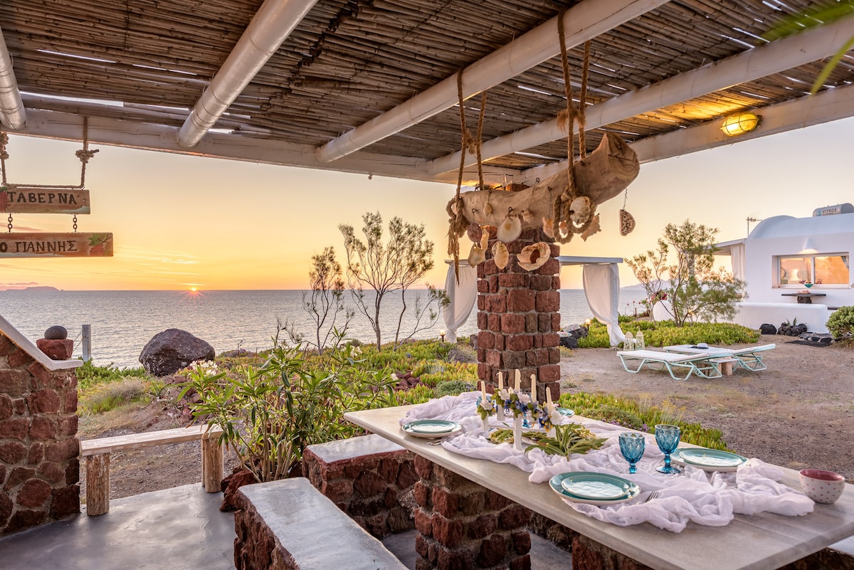 An outdoor dining area features a long table set with plates and glasses. A natural stone structure provides shade, adorned with decorative shells. In the background, the sun sets over the sea, casting a warm glow on the surrounding greenery.